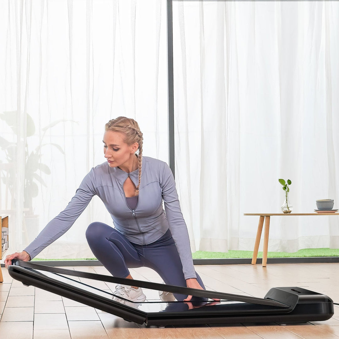 Woman in blue athleisure folding her C2 WalkingPad in living room after exercise, with natural light flooding in the background. 